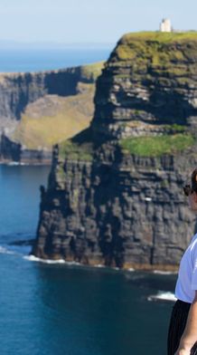 A person with long hair in a bun stands on a cliff, gazing at the ocean and distant cliffs under a clear blue sky.