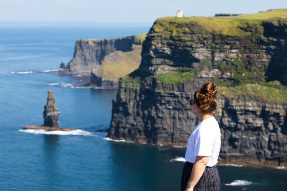 A person with long hair in a bun stands on a cliff, gazing at the ocean and distant cliffs under a clear blue sky.