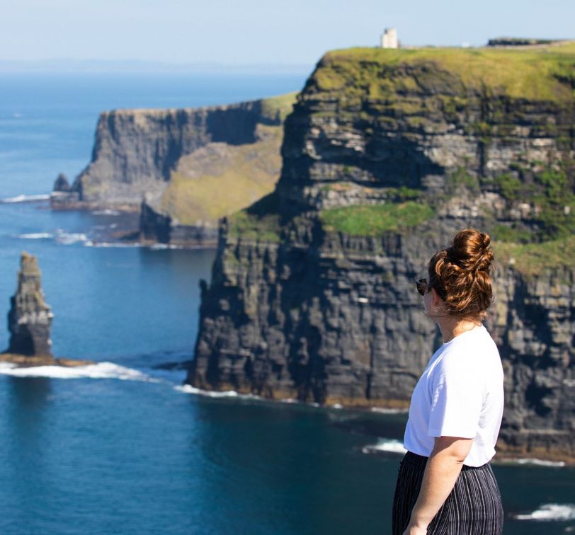 A person with long hair in a bun stands on a cliff, gazing at the ocean and distant cliffs under a clear blue sky.