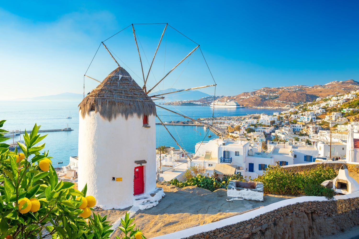 A traditional white windmill with a thatched roof overlooks a vibrant coastal town and sea, with lemons hanging from a nearby tree, under a clear blue sky.