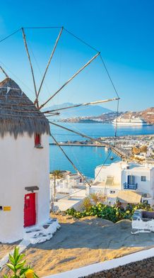 A traditional white windmill with a thatched roof overlooks a vibrant coastal town and sea, with lemons hanging from a nearby tree, under a clear blue sky.