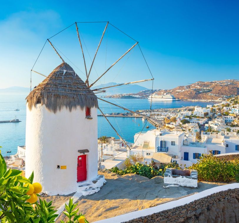 A traditional white windmill with a thatched roof overlooks a vibrant coastal town and sea, with lemons hanging from a nearby tree, under a clear blue sky.