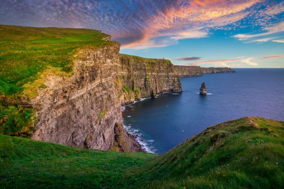 Dramatic cliffs covered in green grass rise above a calm blue sea, with a colorful sunset sky overhead.