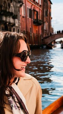 Two women smiling and talking on a boat ride through a picturesque canal, with colorful buildings and a bridge in the background.
