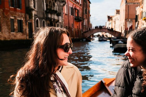 Two women smiling and talking on a boat ride through a picturesque canal, with colorful buildings and a bridge in the background.