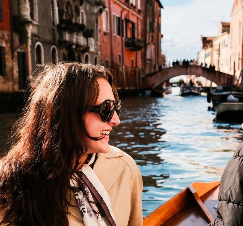 Two women smiling and talking on a boat ride through a picturesque canal, with colorful buildings and a bridge in the background.