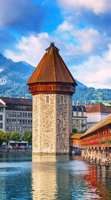 Historic wooden bridge and tower over a river in a European city, surrounded by buildings, with mountain backdrop and a partly cloudy sky.
