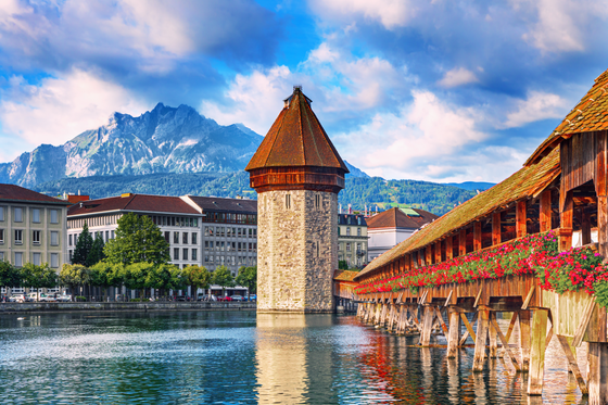 Historic wooden bridge and tower over a river in a European city, surrounded by buildings, with mountain backdrop and a partly cloudy sky.