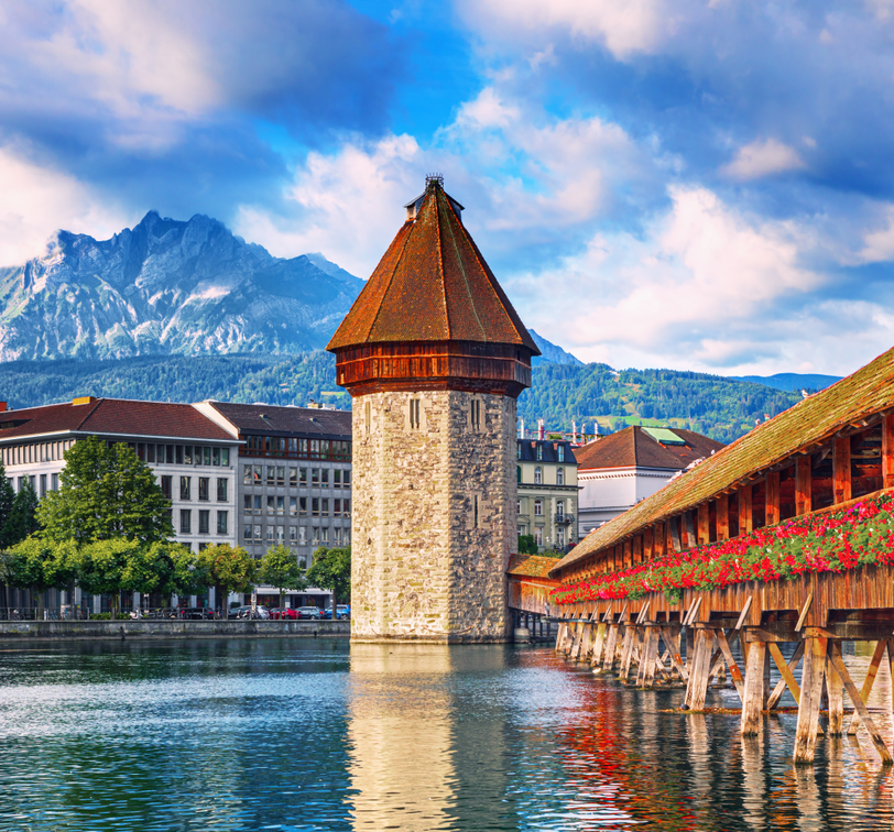 Historic wooden bridge and tower over a river in a European city, surrounded by buildings, with mountain backdrop and a partly cloudy sky.