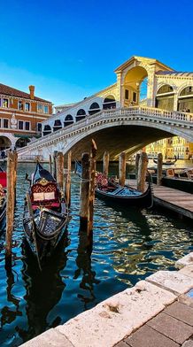 Gondolas moored along a canal beneath the historic Rialto Bridge in Venice, with vibrant buildings in the background under a clear blue sky.