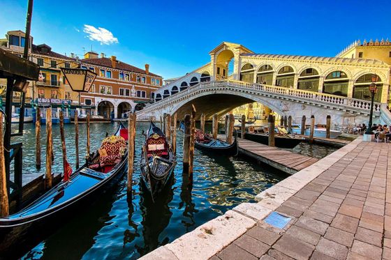 Gondolas moored along a canal beneath the historic Rialto Bridge in Venice, with vibrant buildings in the background under a clear blue sky.