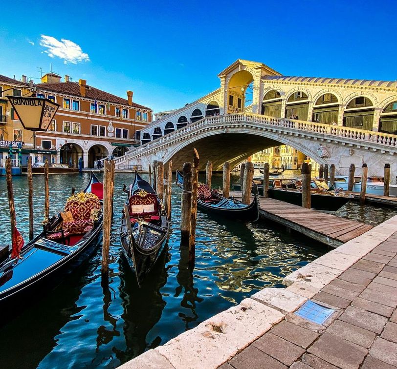 Gondolas moored along a canal beneath the historic Rialto Bridge in Venice, with vibrant buildings in the background under a clear blue sky.
