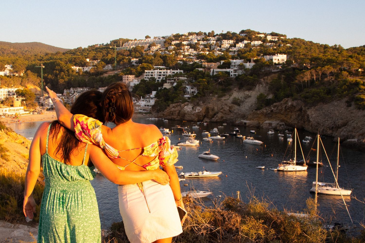Two people in summer dresses stand arm in arm, overlooking a bay with boats and a hillside village at sunset.