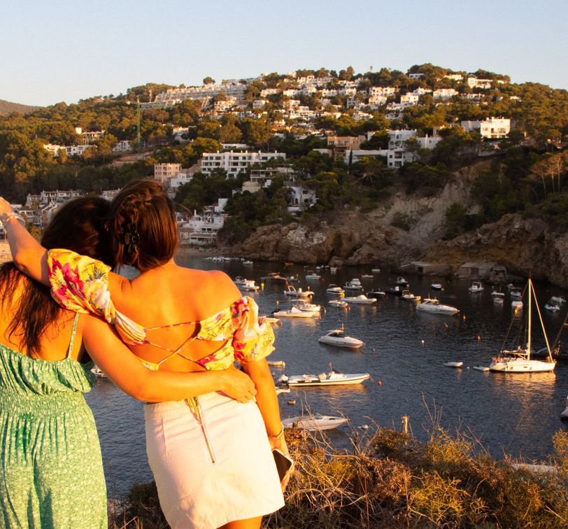 Two people in summer dresses stand arm in arm, overlooking a bay with boats and a hillside village at sunset.