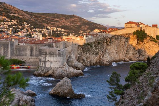 Ancient stone fortress overlooking the sea with a cityscape in the background, rocky cliffs, and lush greenery under a partly cloudy sky.