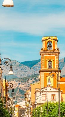 A picturesque street with a bell tower and vintage street lamps, flanked by buildings with balconies, set against a backdrop of mountains.