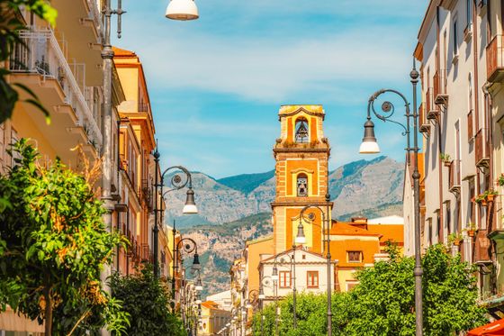 A picturesque street with a bell tower and vintage street lamps, flanked by buildings with balconies, set against a backdrop of mountains.