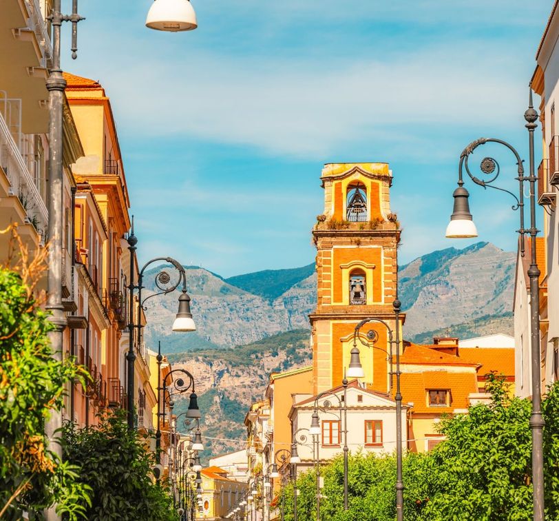 A picturesque street with a bell tower and vintage street lamps, flanked by buildings with balconies, set against a backdrop of mountains.
