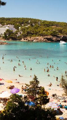 Beach crowded with people enjoying the sun and swimming, colorful umbrellas on the sand, clear turquoise water, and boats anchored in the distance.