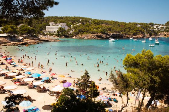 Beach crowded with people enjoying the sun and swimming, colorful umbrellas on the sand, clear turquoise water, and boats anchored in the distance.