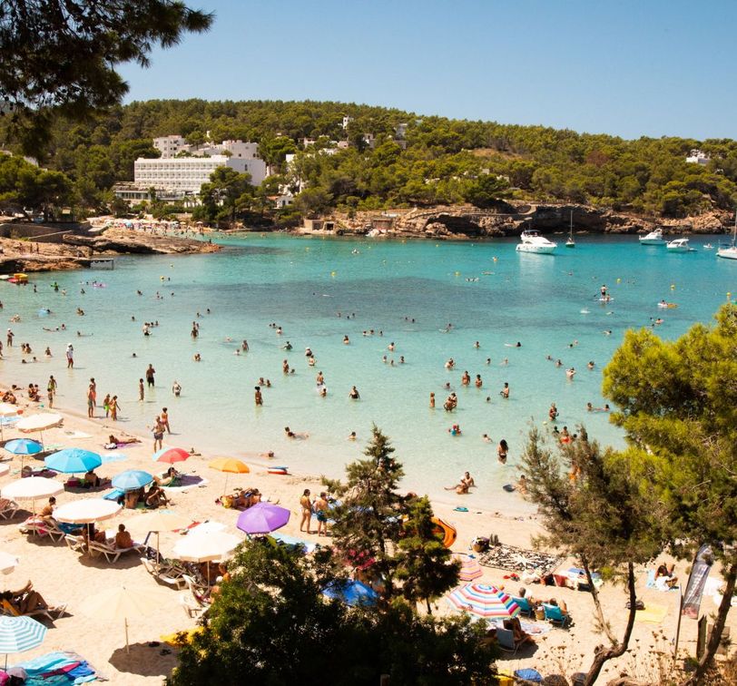 Beach crowded with people enjoying the sun and swimming, colorful umbrellas on the sand, clear turquoise water, and boats anchored in the distance.