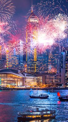Vibrant fireworks illuminate a city skyline at night, with boats on the water reflecting colorful lights from the display and surrounding buildings.