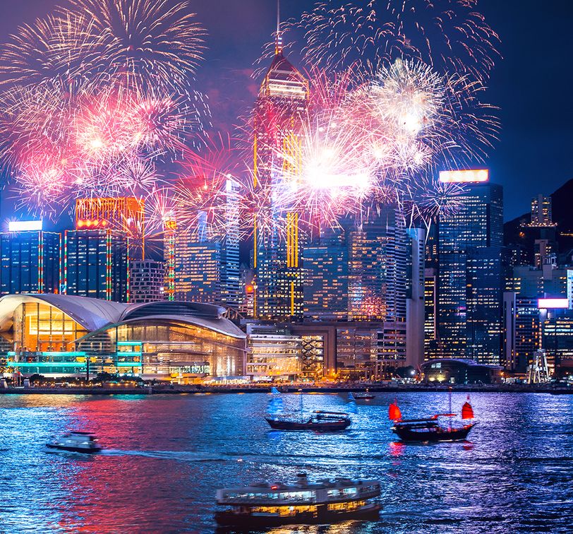 Vibrant fireworks illuminate a city skyline at night, with boats on the water reflecting colorful lights from the display and surrounding buildings.