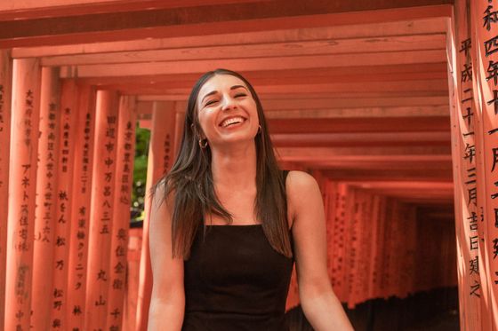 A woman in a black dress smiles brightly while standing in front of red torii gates with inscriptions.