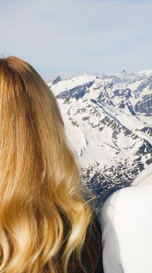 Two women with long hair, one with a loose braid, look out at a panoramic view of snow-covered mountains under a clear sky.