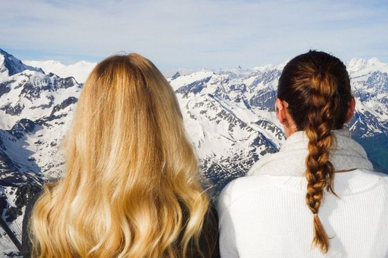 Two women with long hair, one with a loose braid, look out at a panoramic view of snow-covered mountains under a clear sky.