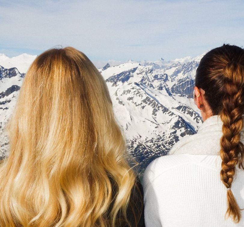 Two women with long hair, one with a loose braid, look out at a panoramic view of snow-covered mountains under a clear sky.