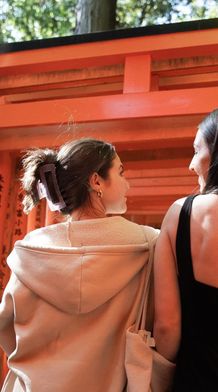 Two women stand side by side in front of vibrant orange torii gates with Japanese inscriptions, surrounded by lush greenery.