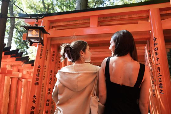 Two women stand side by side in front of vibrant orange torii gates with Japanese inscriptions, surrounded by lush greenery.