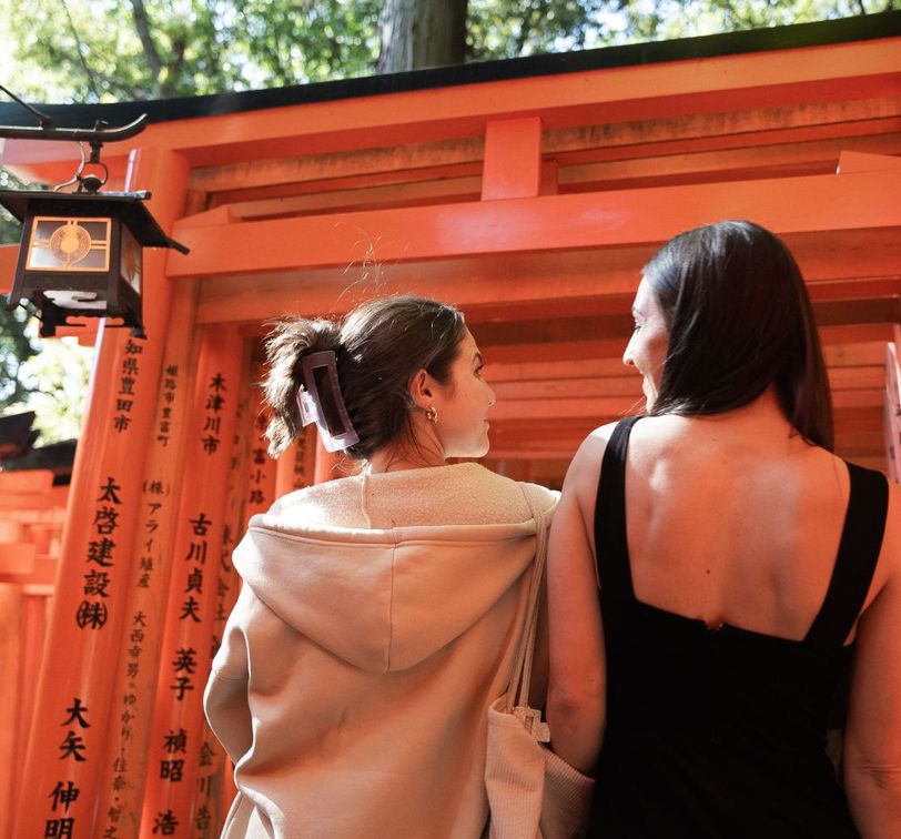 Two women stand side by side in front of vibrant orange torii gates with Japanese inscriptions, surrounded by lush greenery.