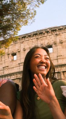 Three people smiling and laughing in front of the Colosseum, with trees and a clear sky in the background.