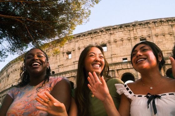 Three people smiling and laughing in front of the Colosseum, with trees and a clear sky in the background.
