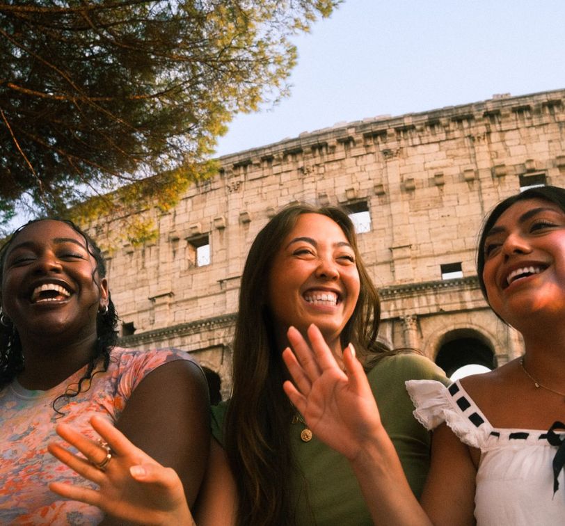 Three people smiling and laughing in front of the Colosseum, with trees and a clear sky in the background.