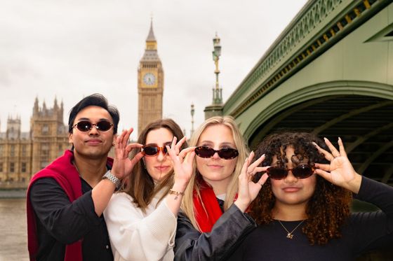 Four people in sunglasses pose playfully in front of Big Ben and a bridge in London.