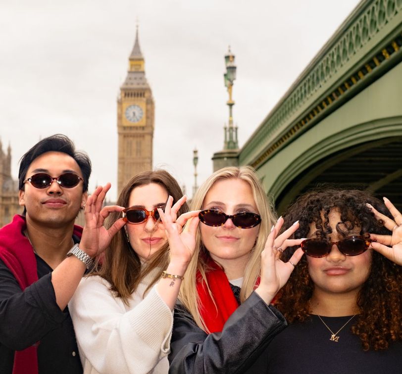 Four people in sunglasses pose playfully in front of Big Ben and a bridge in London.