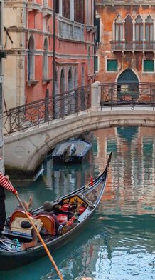 A gondolier navigates a gondola through a narrow, tranquil Venetian canal with colorful, weathered buildings and a stone bridge overhead.