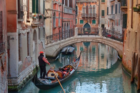 A gondolier navigates a gondola through a narrow, tranquil Venetian canal with colorful, weathered buildings and a stone bridge overhead.