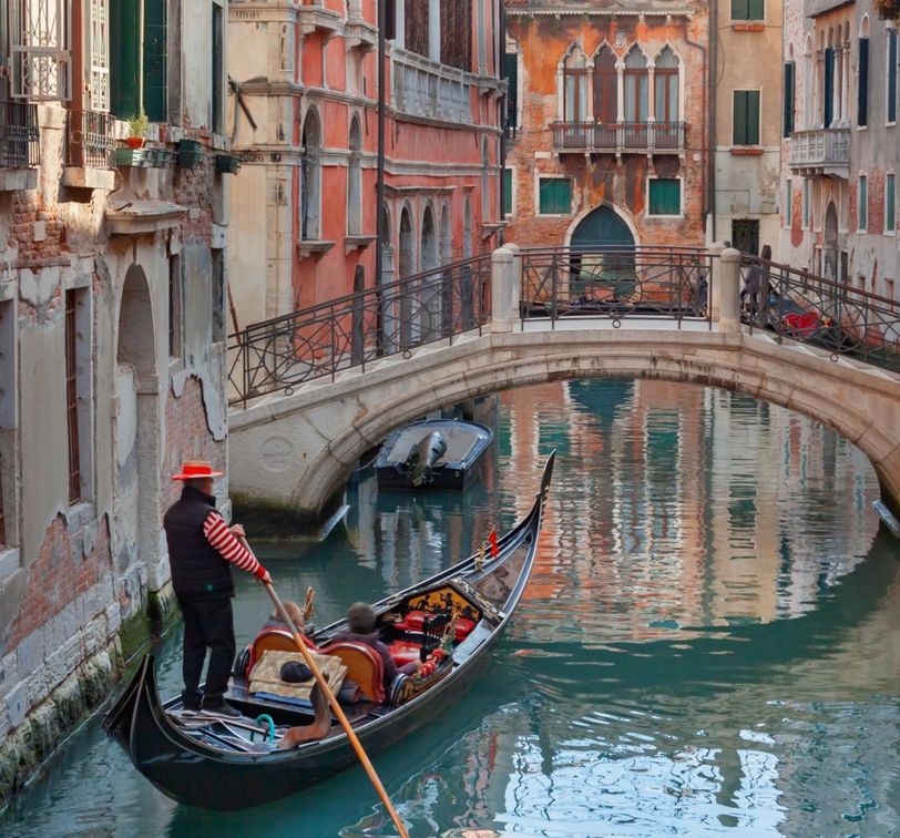 A gondolier navigates a gondola through a narrow, tranquil Venetian canal with colorful, weathered buildings and a stone bridge overhead.