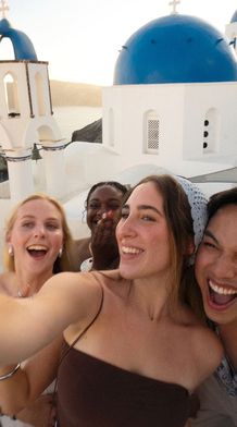 Four friends take a joyful selfie with blue-domed buildings and the sea in the background, capturing a sunny moment in Santorini.
