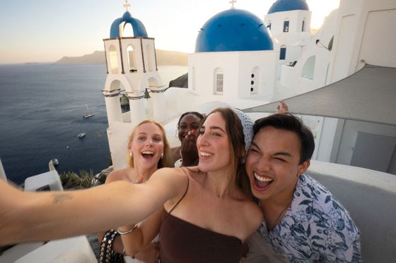 Four friends take a joyful selfie with blue-domed buildings and the sea in the background, capturing a sunny moment in Santorini.