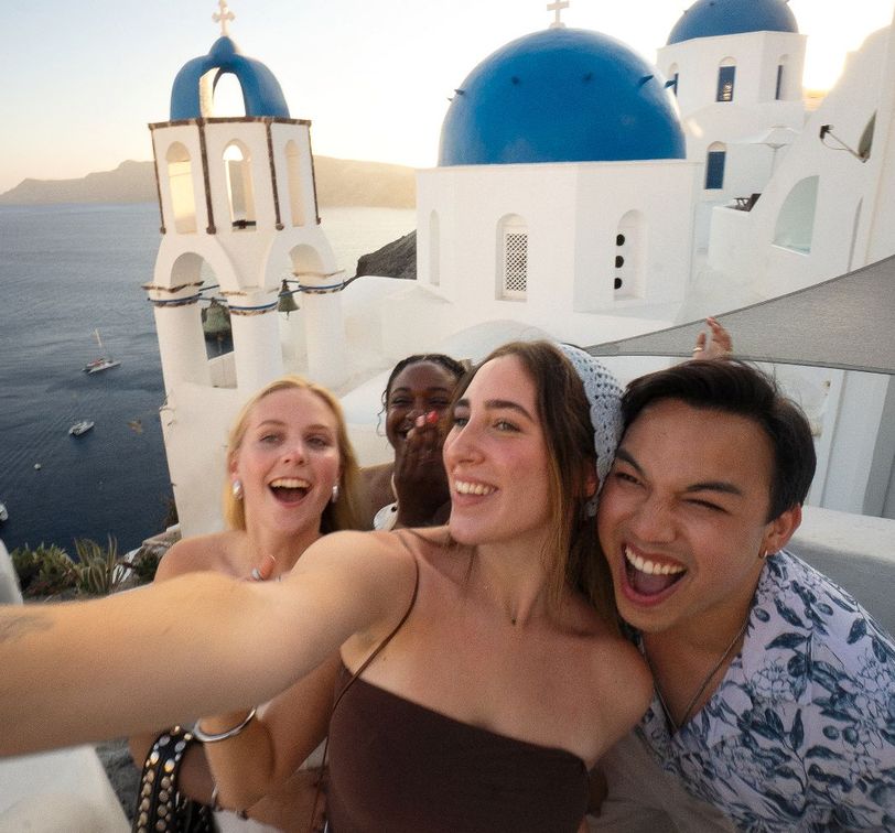 Four friends take a joyful selfie with blue-domed buildings and the sea in the background, capturing a sunny moment in Santorini.