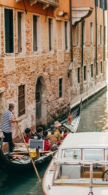 A gondola and a motorboat navigate a narrow canal between brick buildings in Venice, with a gondolier guiding tourists and a man standing by.