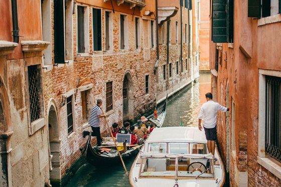 A gondola and a motorboat navigate a narrow canal between brick buildings in Venice, with a gondolier guiding tourists and a man standing by.