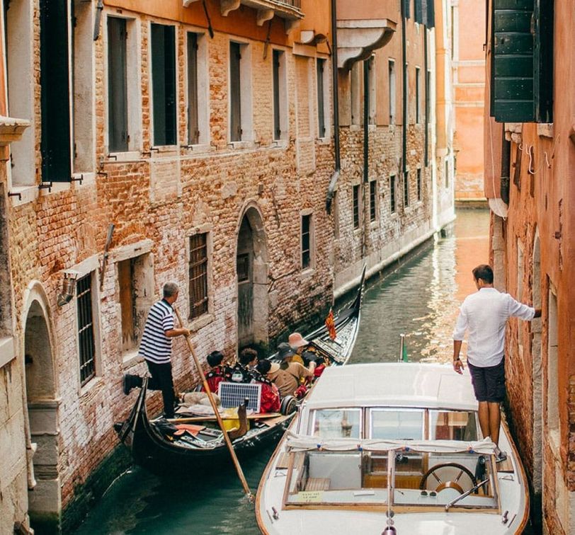 A gondola and a motorboat navigate a narrow canal between brick buildings in Venice, with a gondolier guiding tourists and a man standing by.