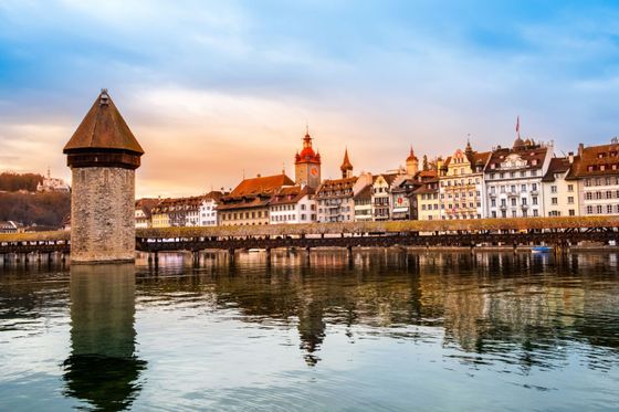 Chapel Bridge and Water Tower in Lucerne at sunset, with colorful historic buildings along the riverbank reflecting in the water.