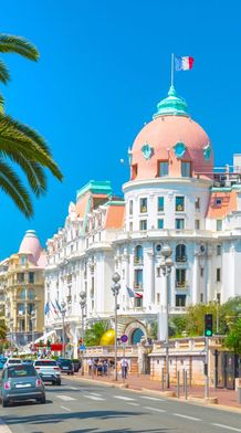 A grand white building with a domed roof and French flag, lined with palm trees on a sunny coastal road, with cars and pedestrians.